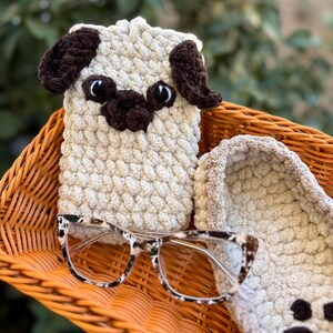 Op de afbeelding: Een handgemaakte, beige gehaakte telefoonhoes voor een mopshond met bruine accenten, in een oranje rieten mand. De mand bevat ook een bril met een schildpadpatroon en een bijpassende gehaakte pantoffel.