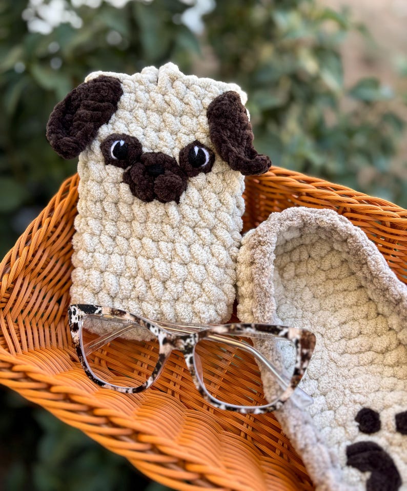 Op de afbeelding: Een handgemaakte, beige gehaakte telefoonhoes voor een mopshond met bruine accenten, in een oranje rieten mand. De mand bevat ook een bril met een schildpadpatroon en een bijpassende gehaakte pantoffel.