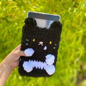 May include: Black and white crochet tablet case shaped like a cat. The case has a black body with white accents, including a bone-shaped design and fluffy white paws. The cat's face features yellow eyes and a pink nose.