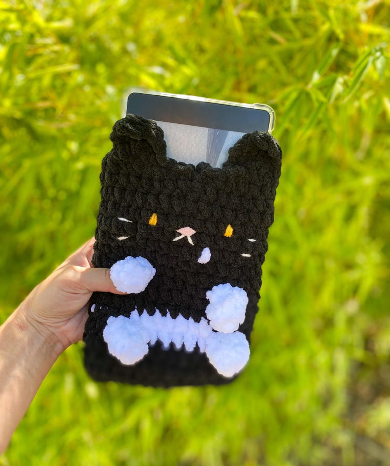 May include: Black and white crochet tablet case shaped like a cat. The case has a black body with white accents, including a bone-shaped design and fluffy white paws. The cat's face features yellow eyes and a pink nose.