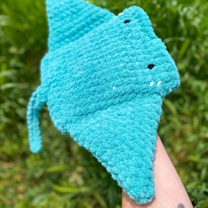 Op de afbeelding: Een gehaakt blauw rog speelgoed met zwarte ogen. Het speelgoed wordt in de hand van een persoon gehouden.