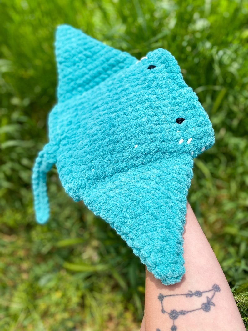 Op de afbeelding: Een gehaakt blauw rog speelgoed met zwarte ogen. Het speelgoed wordt in de hand van een persoon gehouden.