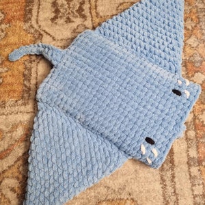 Op de afbeelding: Een blauw gehaakt knuffeldier in de vorm van een mantarog. Het speelgoed is gemaakt van zacht, getextureerd garen en heeft een witte gehaakte mond.