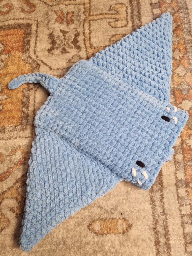 Op de afbeelding: Een blauw gehaakt knuffeldier in de vorm van een mantarog. Het speelgoed is gemaakt van zacht, getextureerd garen en heeft een witte gehaakte mond.