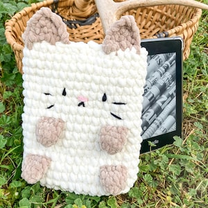 May include: A white, crocheted cat-shaped Kindle cover with tan ears and paws. The cover has black stitched eyes and whiskers, and a pink nose. The Kindle is visible inside the cover, resting on a bed of grass.