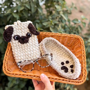 Op de afbeelding: Een beige gehaakte telefoonhoes in de vorm van een mopshond, een bijpassende slipper met een pootafdruk en een bril met een schildpadpatroon liggen in een rieten mand. De items zijn handgemaakt en zien er gezellig uit.