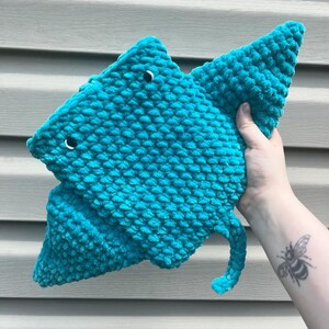 Op de afbeelding: Een tealblauw gebreid knuffeldier in de vorm van een mantarog met zwarte ogen. Het speelgoed wordt vastgehouden door een hand met een bijentattoo.