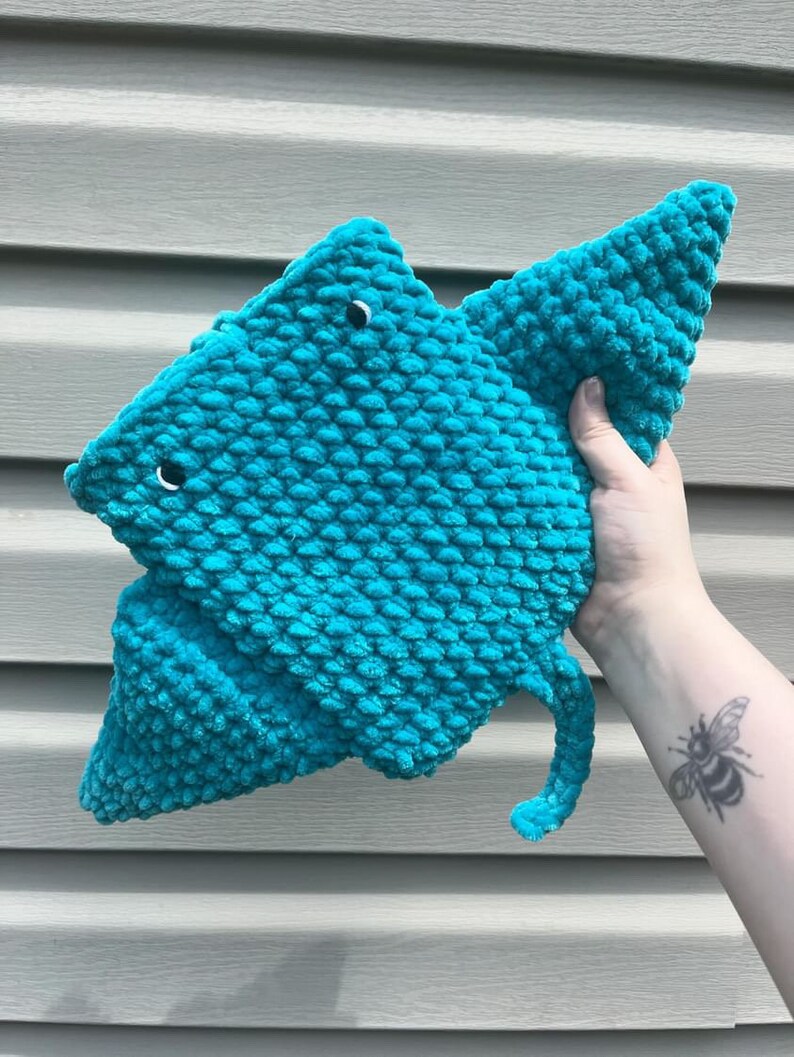 Op de afbeelding: Een tealblauw gebreid knuffeldier in de vorm van een mantarog met zwarte ogen. Het speelgoed wordt vastgehouden door een hand met een bijentattoo.