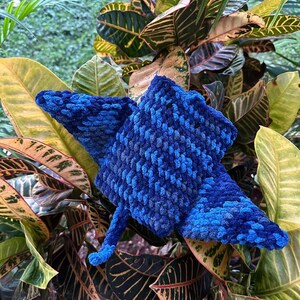 Op de afbeelding: Een blauw gehaakt stervormig pluche speelgoed met een textuur. Het speelgoed ligt op een bed van groene en gele bladeren.