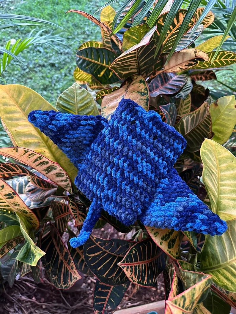 Op de afbeelding: Een blauw gehaakt stervormig pluche speelgoed met een textuur. Het speelgoed ligt op een bed van groene en gele bladeren.