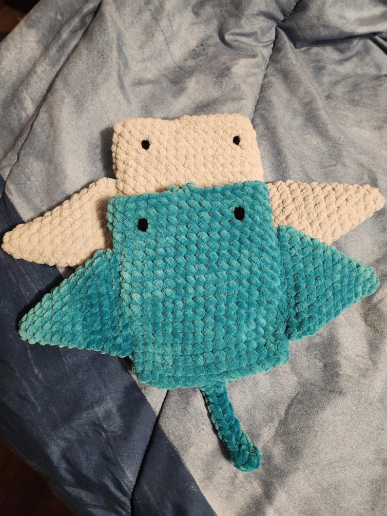 Op de afbeelding: Een gehaakt knuffeldier in de vorm van een rog in tealblauw en beige met zwarte ogen. De rog heeft een lange staart en twee grote vinnen.