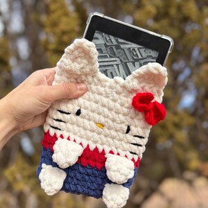 May include: A white, red, and blue crochet Kindle case shaped like a cat. The case has black eyes, a yellow nose, and a red bow. The case is designed to hold an e-reader.