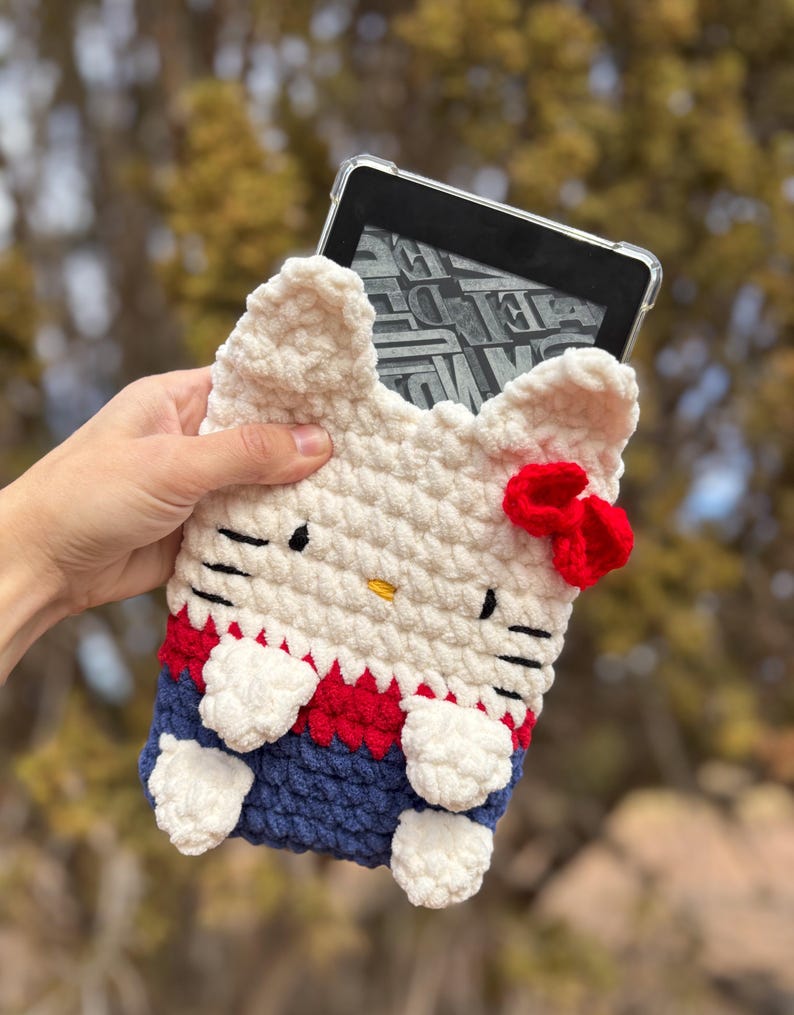 May include: A white, red, and blue crochet Kindle case shaped like a cat. The case has black eyes, a yellow nose, and a red bow. The case is designed to hold an e-reader.