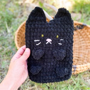 May include: A black crochet cat-shaped pouch with white whiskers, a pink nose, and yellow eyes. The pouch has two small ears and two heart-shaped paws. The item is handmade and appears to be soft and plush.