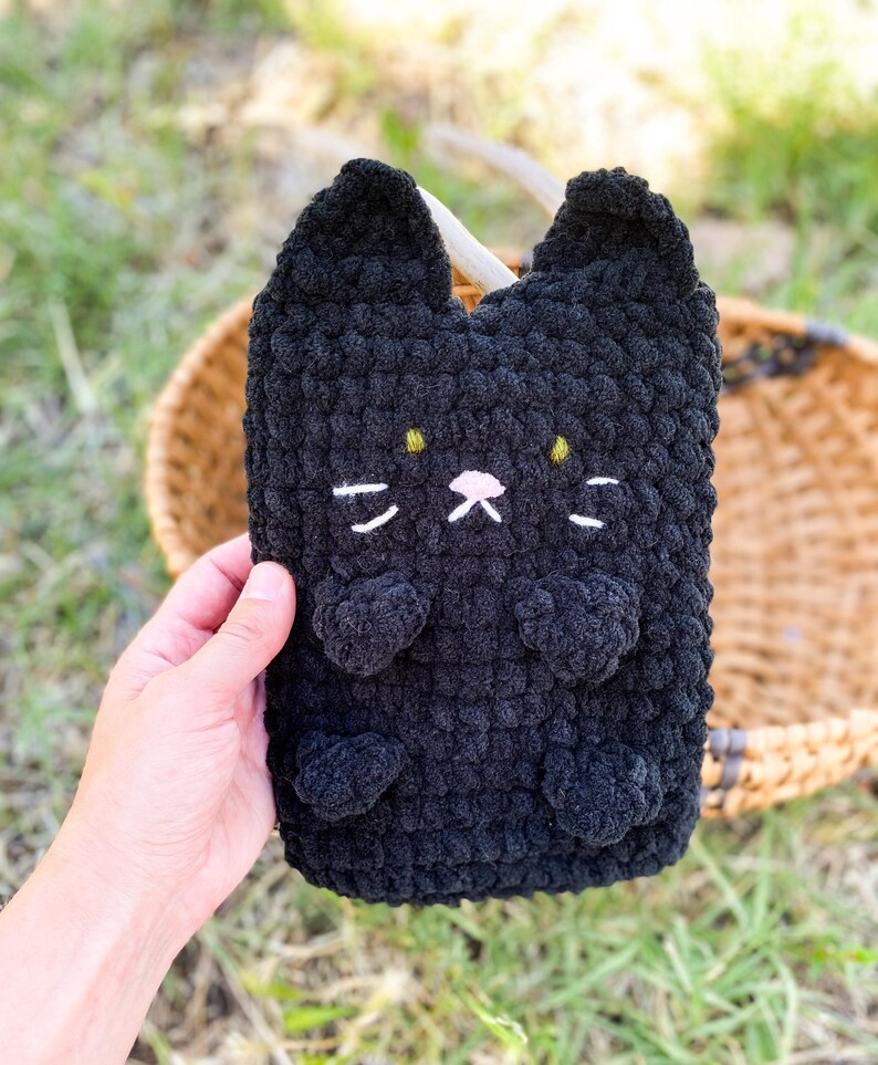 May include: A black crochet cat-shaped pouch with white whiskers, a pink nose, and yellow eyes. The pouch has two small ears and two heart-shaped paws. The item is handmade and appears to be soft and plush.