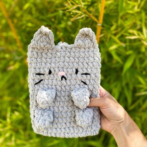 May include: A gray crocheted cat-shaped item with black whiskers, a pink nose, and small ears. The item has two small paws and is made of a soft, textured yarn. The background is blurred green foliage.
