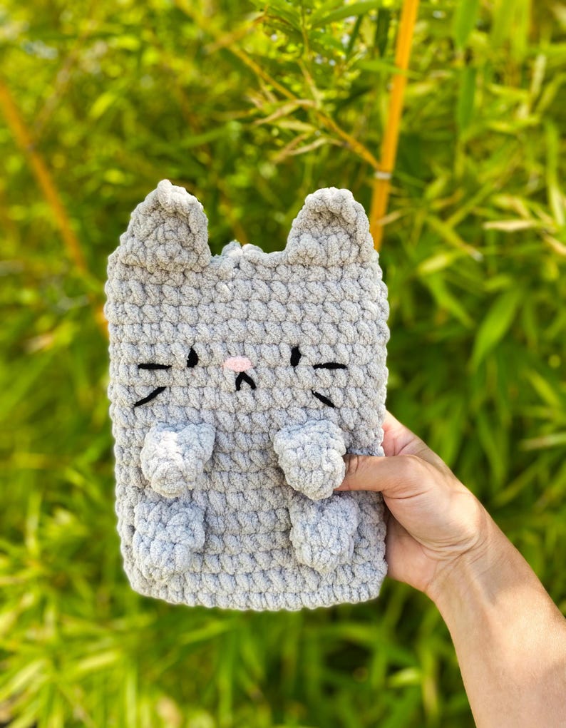 May include: A gray crocheted cat-shaped item with black whiskers, a pink nose, and small ears. The item has two small paws and is made of a soft, textured yarn. The background is blurred green foliage.