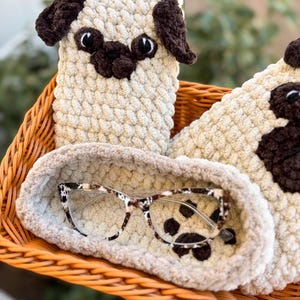 Op de afbeelding: Een beige en bruin gehaakte brillenkoker in de vorm van een mopshond en een bijpassende brillenhoes. De brillenkoker bevat een bril met een schildpadpatroon. De items liggen in een gevlochten mand.
