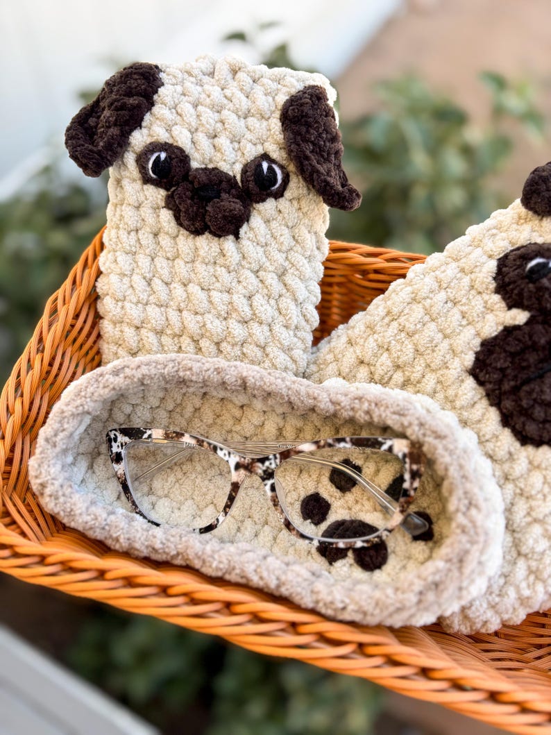 Op de afbeelding: Een beige en bruin gehaakte brillenkoker in de vorm van een mopshond en een bijpassende brillenhoes. De brillenkoker bevat een bril met een schildpadpatroon. De items liggen in een gevlochten mand.