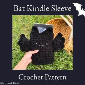 Op de afbeelding: Zwarte gebreide Kindle-hoes in de vorm van een vleermuis met een schattig gezicht. De hoes ligt op een bed van groen gras. Haakpatroon.