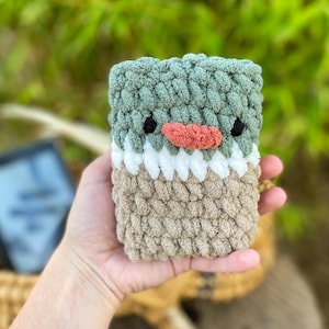 May include: A handmade, crochet phone case in the shape of a bird. The case features a sage green top, white middle, and tan bottom. It has black eyes and a coral-colored beak. The case is held in a hand.