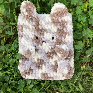 May include: A handmade, plush cat-shaped pillow in shades of brown, white, and gray. The pillow features a pink nose, black eyes, and small, rounded ears. The soft, textured material gives the pillow a cozy appearance.