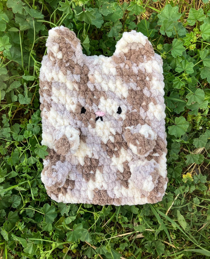 May include: A handmade, plush cat-shaped pillow in shades of brown, white, and gray. The pillow features a pink nose, black eyes, and small, rounded ears. The soft, textured material gives the pillow a cozy appearance.