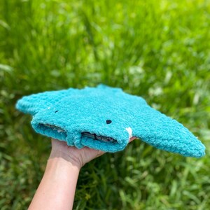 Op de afbeelding: Een tealblauw gebreid knuffeldier in de vorm van een mantarog met zwarte ogen. Het speelgoed wordt in een hand gehouden met een groene grasachtige achtergrond.