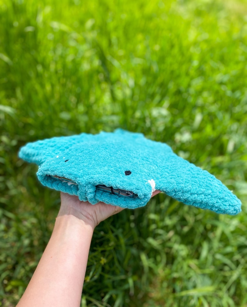 Op de afbeelding: Een tealblauw gebreid knuffeldier in de vorm van een mantarog met zwarte ogen. Het speelgoed wordt in een hand gehouden met een groene grasachtige achtergrond.