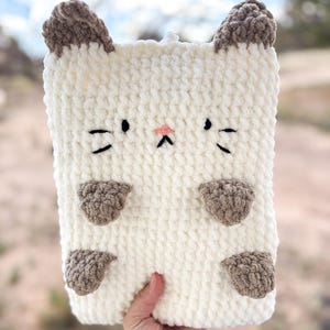 May include: A cream-colored, rectangular crochet cat-shaped case with brown ears, paws, and facial details. The cat face features black eyes and whiskers, and a pink nose. The case is held by a hand.