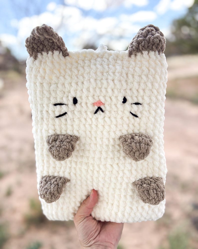 May include: A cream-colored, rectangular crochet cat-shaped case with brown ears, paws, and facial details. The cat face features black eyes and whiskers, and a pink nose. The case is held by a hand.