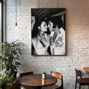 Pode incluir: Impressão emoldurada em preto e branco de um casal em um abraço íntimo. O homem usa uma camisa branca com bordados ornamentados, e a mulher uma blusa branca. A impressão está pendurada em uma parede de tijolos brancos acima de uma mesa redonda de madeira e cadeiras.