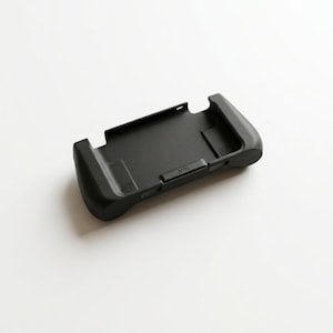 May include: A black handheld gaming console grip, designed to enhance comfort and control. The grip features ergonomic handles and a central slot for the console. The device is made of matte black plastic and is shown on a white background.