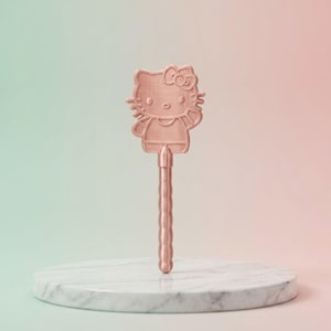 Puede incluir: Un bolígrafo en forma de Hello Kitty de color cobre, de pie sobre un pedestal de mármol blanco. El bolígrafo presenta un contorno detallado del personaje icónico, con un lazo y rasgos faciales. El mango del bolígrafo tiene un diseño acanalado. El fondo es un degradado de verde a rosa.