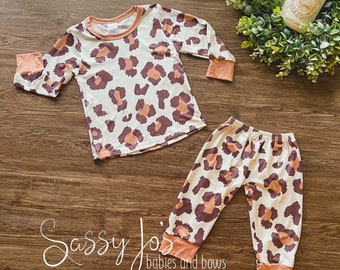 Peut inclure: Un ensemble pyjama deux pièces pour enfant, composé d'un haut à manches longues et d'un pantalon à imprimé léopard brun et pêche sur fond blanc. L'ensemble est doté d'une bordure pêche autour du col, des poignets et des jambes du pantalon. Le texte "Sassy Jo's babies and bows" est visible sur l'image.