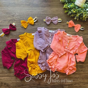 May include: Four baby onesies in different colors: burgundy, mustard yellow, lavender, and peach. Each onesie has long sleeves, a ruffled neckline, and a button closure. The onesies are arranged in a semi-circle on a wooden surface.  There are four matching bows in the same colors as the onesies.