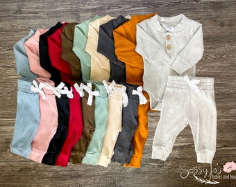 Peut inclure: Un ensemble de 10 bodys bébé à manches longues côtelés et pantalons assortis dans différentes couleurs, dont gris, rouge, noir, marron, beige et orange. Les bodys ont une fermeture boutonnée au niveau du cou. Les pantalons ont une taille à cordon. "Sassy Jo's Babies and Bows" est imprimé dans le coin inférieur droit de l'image.