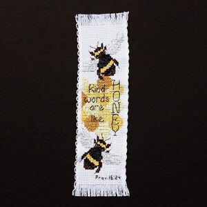 May include: A white cross-stitch bookmark with fringed edges. The design features two bees, honeycombs, and the text "Kind words are like honey" with the Bible verse Proverbs 16:24.