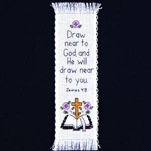 May include: A white, cross-stitched bookmark with the text "Draw near to God, and He will draw near to you. James 4:8." The bookmark features a cross, an open book, and floral accents in purple and orange.