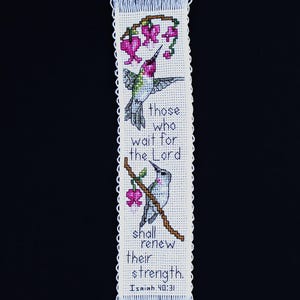 May include: A rectangular white fabric bookmark featuring cross-stitch embroidery of two hummingbirds and pink flowers on brown branches. The text reads: those who wait for the Lord shall renew their strength. Isaiah 40:31. It has fringed edges.