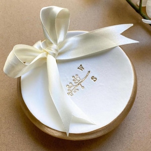 May include: A white ceramic dish with a gold rim, tied with a cream satin ribbon bow. The dish features gold embroidered initials "W" and "S" with a floral branch design. The overall aesthetic is elegant and decorative.