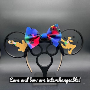 Puede incluir: Un par de orejas de ratón negras con un perro de dibujos animados marrón y tostado en cada oreja. Un lazo colorido con huellas de patas está unido a la diadema. El texto "Ears and bow are interchangeable!" está en la parte inferior de la imagen.