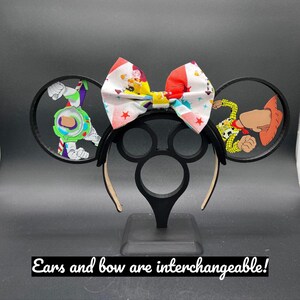 May include: Black mouse ears headband with a red, white, and blue bow. The ears feature a clear plastic overlay with images of Woody and Buzz Lightyear from Toy Story. The text "Ears and bow are interchangeable!" is visible at the bottom of the image.