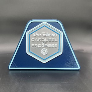 May include: A blue and light blue triangular sign featuring the text "Walt Disney's Carousel of Progress" in silver lettering. The sign has a hexagonal emblem in the center with a circular design. The sign is a decorative item.