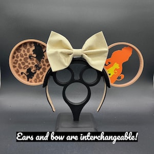 May include: A pair of brown mouse ears with a beige bow. The ears feature a silhouette of a lion and a leopard print. The text "Ears and bow are interchangeable!" is visible at the bottom of the image.