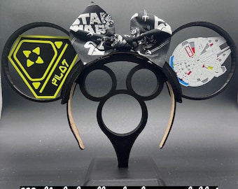 Puede incluir: Diadema negra de orejas de ratón con un lazo temático de Star Wars y dos decoraciones circulares. Una decoración presenta un Halcón Milenario y la otra un letrero de advertencia amarillo y negro con el texto "PILOT 7".