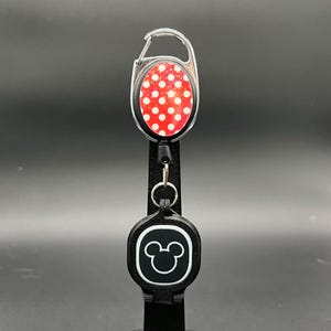 May include: A retractable badge reel with a red and white polka dot design. The reel has a black backing and a silver carabiner clip. The bottom of the reel features a black square with a white Mickey Mouse head.