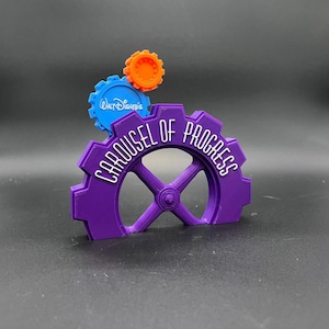 May include: A purple gear-shaped sculpture with the words "Carousel of Progress" in white. Above it are two smaller gears, one blue with "Walt Disney's" and the other orange. The sculpture is on a dark surface.