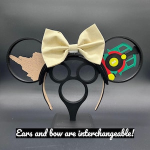 May include: A pair of black mouse ears with a beige bow. One ear features a brown silhouette of a castle, the other ear features a green, red, and yellow silhouette of a train. The text "Ears and bow are interchangeable!" is at the bottom of the image.