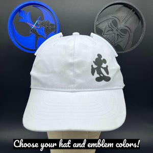 Puede incluir: Gorra de b&eacute;isbol blanca con una silueta negra de Mickey Mouse. La gorra tiene dos accesorios en forma de oreja y se muestra con dos emblemas circulares, uno azul con un dise&ntilde;o de Star Wars y otro negro con la cara de Darth Vader. Texto: "&iexcl;Elige los colores de tu sombrero y emblema!"